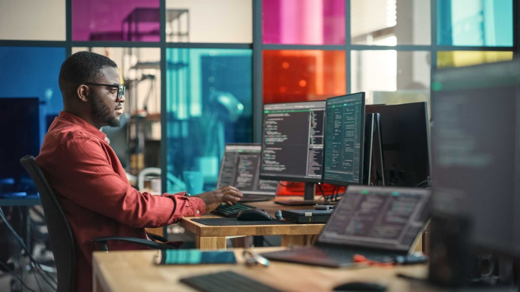Automated Software Update Services. A man sits in a colorful tech office, focused on coding across multiple monitors in a modern, vibrant workspace.